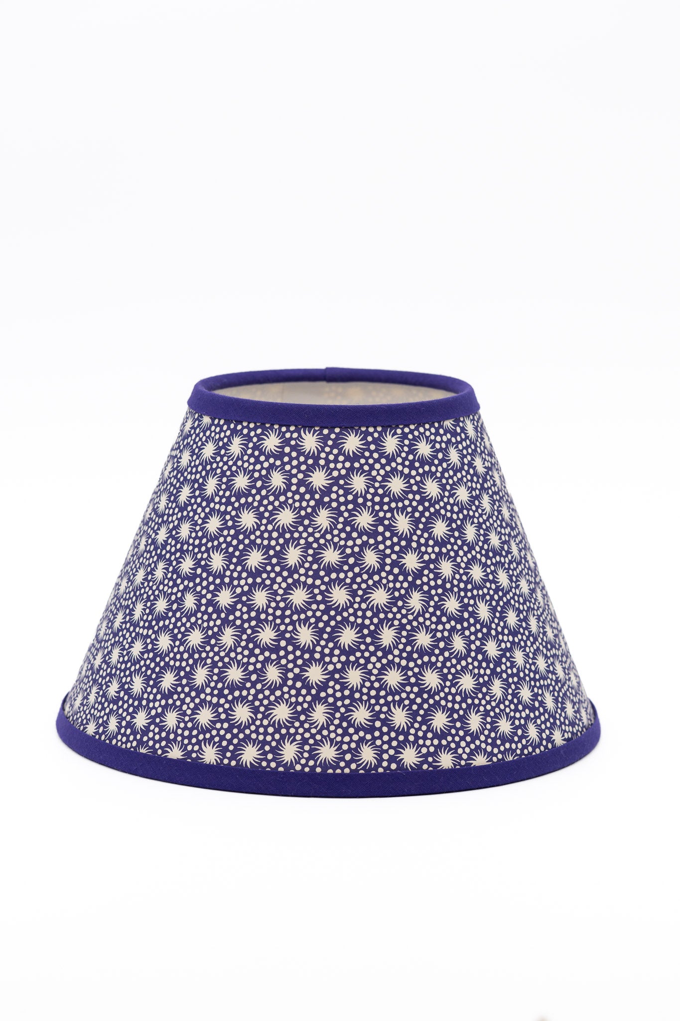 A paper lampshade with a repeating white sunburst pattern on a deep purple background, finished with solid purple trim at the top and bottom.