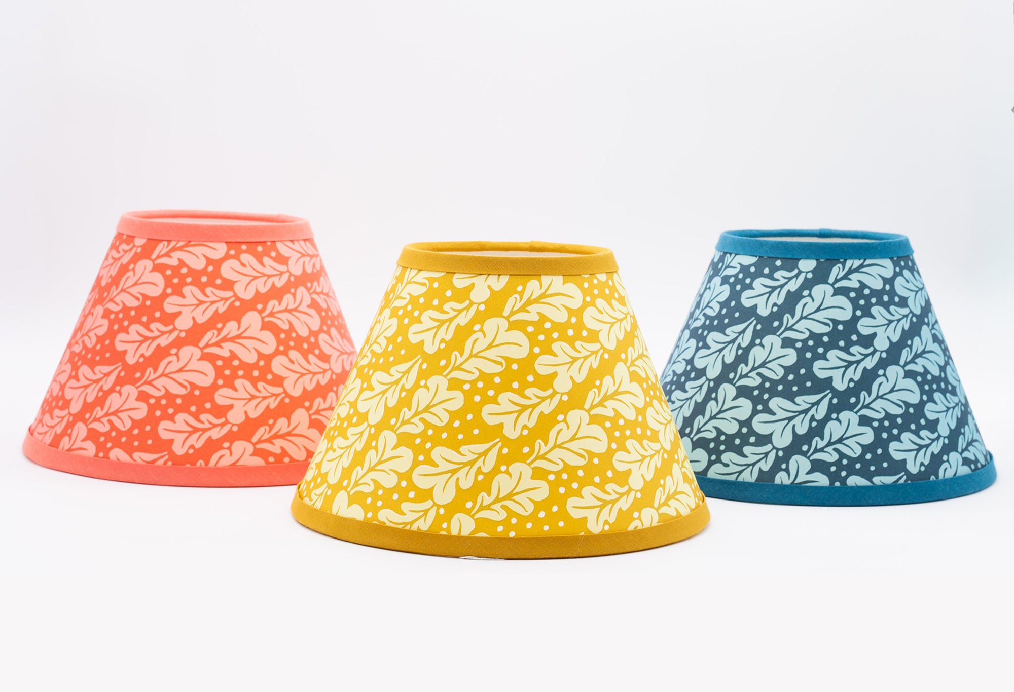Three fabric lampshades with colourful leaf patterns, shown in coral orange, mustard yellow, and teal blue on a white background.