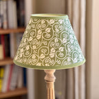 A table lamp with a green and cream floral patterned paper shade and a light wood base, set against a softly blurred background of a bookshelf and curtains.