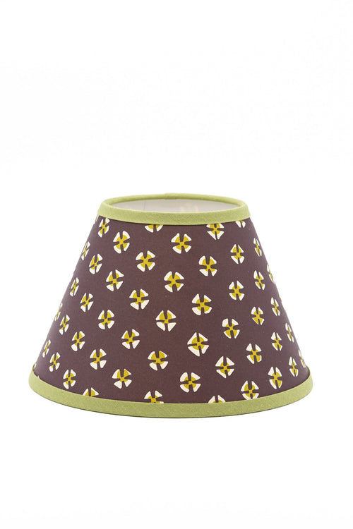 A paper lampshade with a dark brown background, decorated with a repeating pattern of yellow and white stylized crosses, finished with light green trim at the top and bottom.