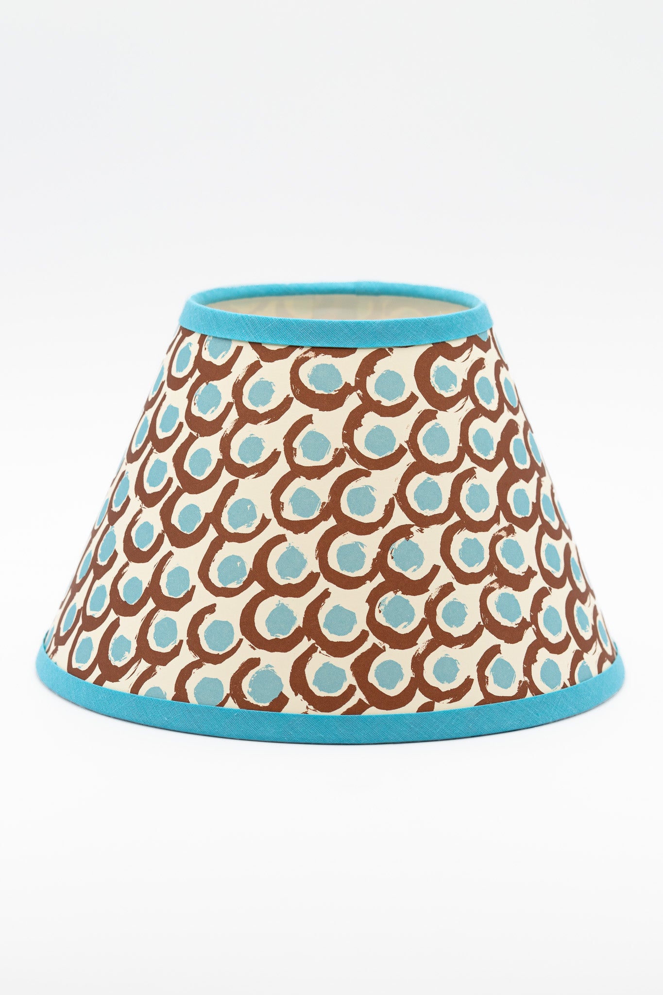 A paper lampshade with a repeating brown and light blue circular pattern on a cream background, finished with a solid turquoise blue trim at the top and bottom.