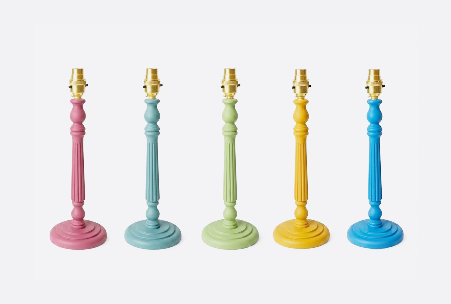 Five pastel-colored table lamps in pink, light blue, mint green, mustard yellow, and royal blue, arranged in a row against a light gray background.