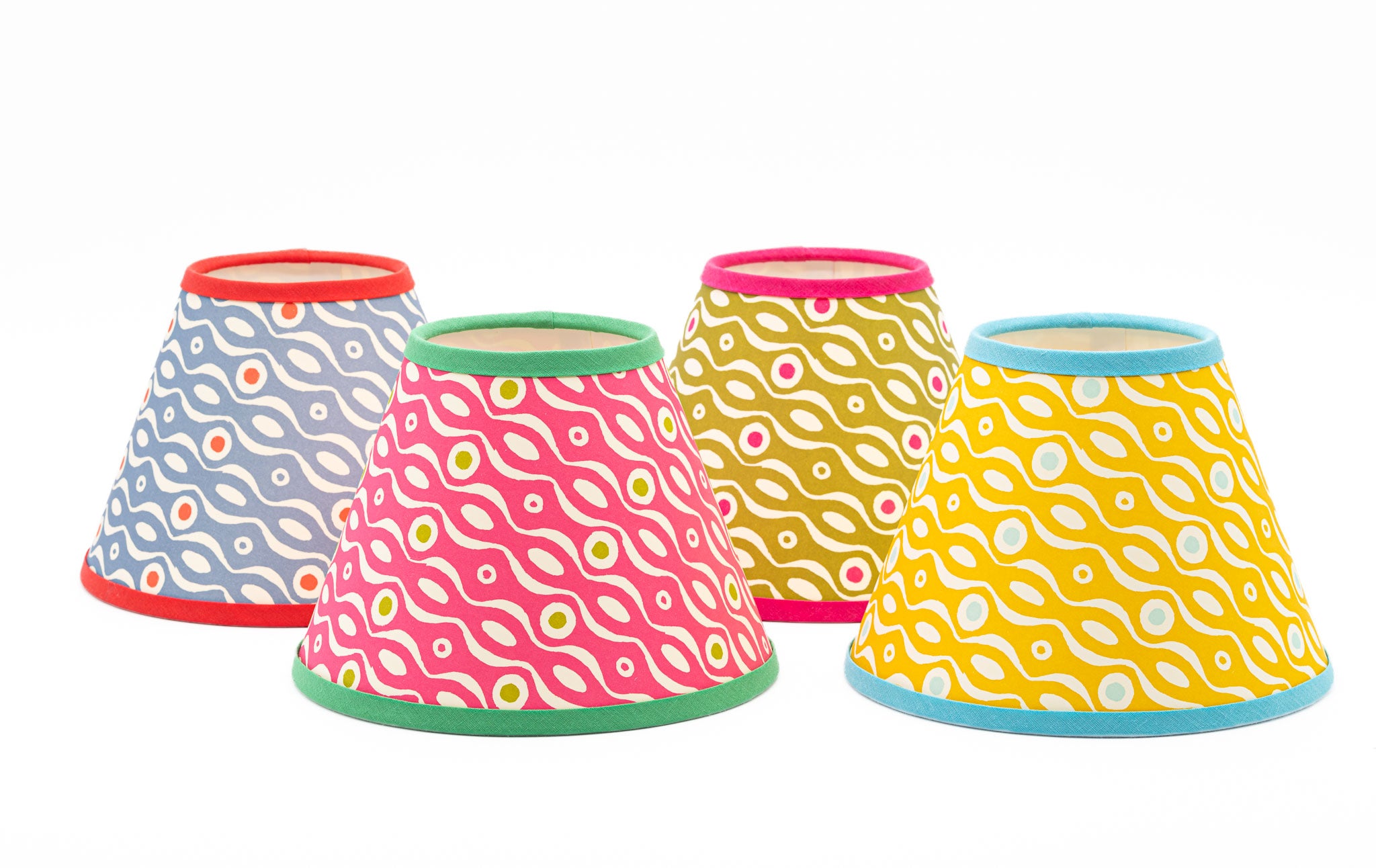 Four fabric lamp shades with colorful wavy dot patterns, arranged in a row against a plain white background. The shades feature different color combinations: blue-gray, pink, mustard yellow, and bright yellow, each with contrasting trim at the top and bottom.
